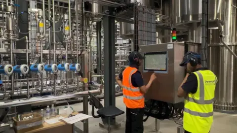 Two people in caps, wearing headphones and high-vis jackets, one in orange, one in yellow/green, stand next to a big metal box, their backs to us. The box is connected to lots of other metal machinery behind them in this factory.
