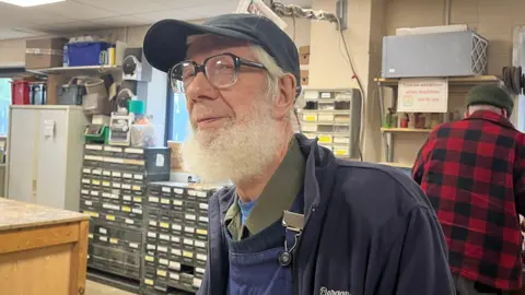 Isabella Verona/BBC A close-up of an older man wearing a black cap and glasses, standing in a workshop. The person has a white beard and is wearing a dark jacket. Behind him are shelves filled with small drawers, tools, and containers. In the background, another person in a red-and-black checkered shirt stands facing away.