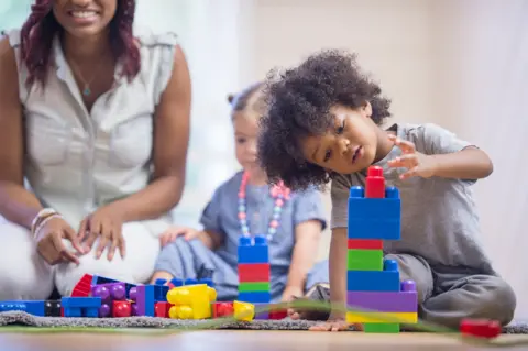 Getty Images A stock image showing a young boy in the foreground playing with large stackable lego-type bricks. A young girl and a woman can be seen in the background also on the playmat.