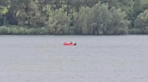BBC A red rescue boat with a person in it on a reservoir. Trees are seen behind the water.