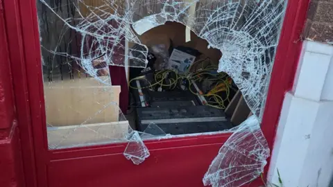 Jowayne Marks A window set inside a red frame is smashed with a large hole. Inside you can see wires and a toolbox