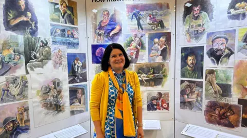 Laura Joint Louise Pasterfield stands in front of an exhibition of her watercolour paintings which depict people in Ukraine under the Russian invasion 
