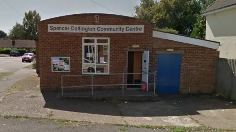 Google A brick building, with a blue door to the right, and an open white door. There are also two windows and a sign on the building. A car is parked to the left and there is a ramp and a metal railing in front of the building. 