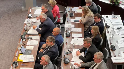 Two rows of councillors sit on a table, with papers and laptops in front of them. They are all looking towards the front of the room. 
