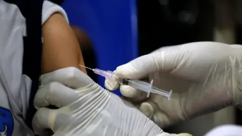 A person wearing white medical gloves injects a needle containing a clear, colourless vaccine into a child's arm. 