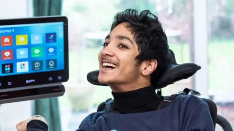 Image of a young man with black hair in a wheelchair, smiling at the camera. In the background can be seen a tablet screen and large French doors on to a garden.