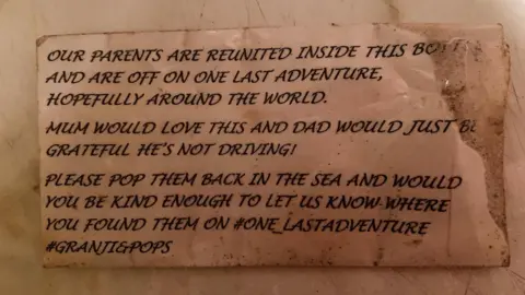 Mike Nicholls A piece of paper that says "Our parents are reunited inside this bottle and are off on one last adventure, hopefully around the world. Mum would love this and Dad would just be grateful he's not driving! Please pop them back in the sea and would you be kind enough to let us know where you found them on #one_lastadventure #Granji&Pops."
