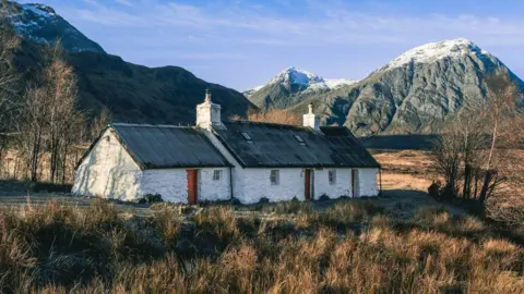 John Thomson A white cottage with a dark roof. There are two buildings with three doors. The building closest to the camera is slightly smaller. It has an orange door in the middle and a small window in the top right of the building. The larger building also has a dark roof and has two doors and two small windows. A large hill is in the background and is dark coloured with white snow on the top. The cottage is surrounded by long, green and yellow grass.