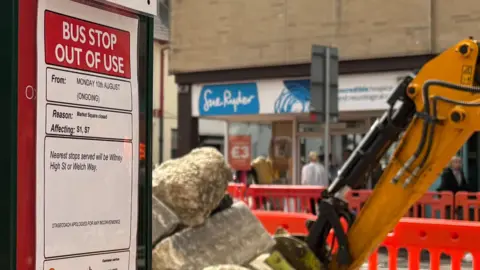 A closed bus stop with rubble and and a digger in the background and orange fencing around it.