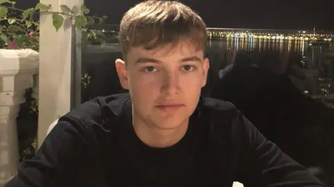 Jon Smith Seb, a teenage boy, looking at the camera. He has a neutral expression. He has straight black hair which is short on the sides and falls over his forehead. He is in a long-sleeved black shirt. He appears to be sat at a table on holiday, with a marina behind him at night.