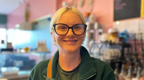 George Carden/BBC Rachel Warren with coffee syrups behind her in her cafe and a slushy machine, she's wearing a green apron, has blonde hair and glasses