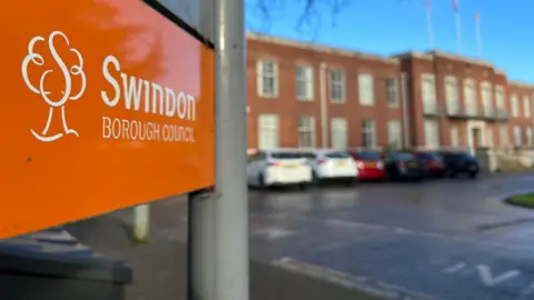 A shot of a sign outside Swindon Borough Council's offices with the council's logo on an orange background. The building is int he background and out of focus