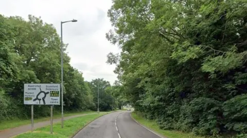 Jubilee Avenue is a road surrounded by trees and shrubs. There is a footpath to the left of the image. There is also a road sign pointing to nearby routes to places in the county and beyond