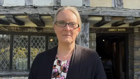 A woman with blonde hair, glasses, a flowery top and a grey cardigan stood outside a building with a lead-framed window