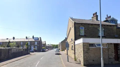 A residential road with the gable end of a terrace of houses on the right hand side. On the left, further away, a row of terraced houses. The sky is blue. There are some cars parked alongside the street.
