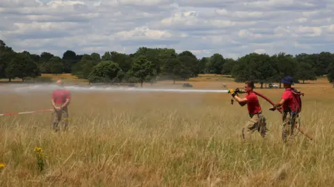 Two firefighters holding hose blast water from a hosepipe as a colleague watches on. All are wearing read and standing in the long grass at a cloudy Richmond Park