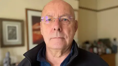 Simon Thake A bald man with glasses and wearing a black fleece jacket stands in a front room