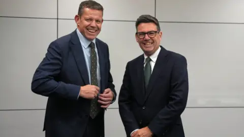 PA Media Rhun ap Iorwerth and Andy Burnham in dark suits and green ties stand side by side in front of a plain grey panelled wall during a formal indoor photo opportunity. They are both laughing.