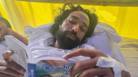 Devendra Nath Mahto laying on a mattress wearing white clothes and reading a book 