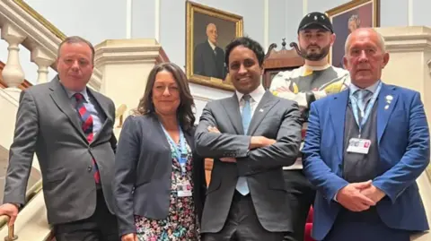 Five people stood on a grand staircase. Four are in business attire and one man is wearing casual clothes. They are Arron Banks, Linden Kemkaran, Zia Yusuf, Nathaniel Fried and Brian Collins. 