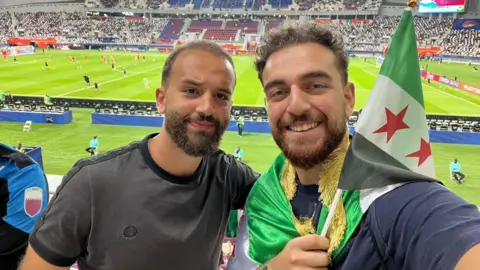 Bader Al-Akkad Bader and Amer Al-Akkad take a selfie in the stands at a football stadium, with Amer holding a Syrian flag.