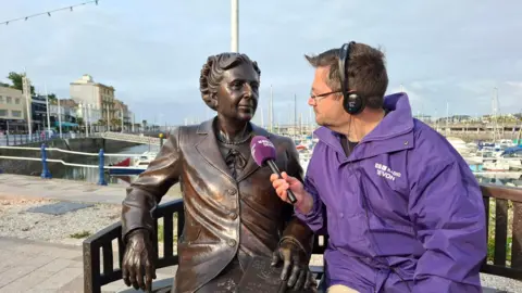 A bronze statue of Agatha Christie, sat on a bench with a man to her left. The man is wearing a BBC Radio Devon purple coat and his holding a microphone mimicking talking with the statue. Behind them is the Torquay marina.