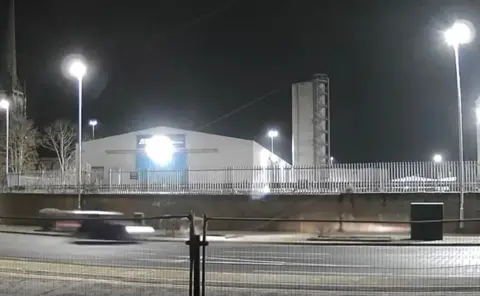 A still photo of a car in motion driving along a road near to a telecoms cabinet. It is night time and it is the only car on the road.