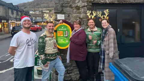 Tina Salway stands with three male volunteers in front of a green and yellow defibrillator that's been installed on a stone wall outside a business. 