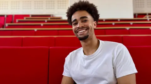 Hazel Shearing / BBC Kieran McRae is in a white T-shirt seated in a lecture theatre with rows of bright red tiered seats. The image is framed as a portrait, with the seating stretching into the background of the auditorium.