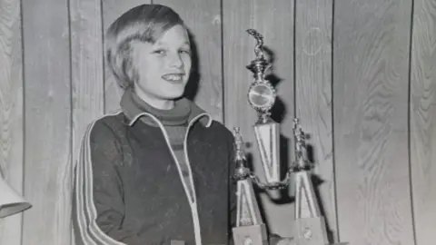 Adam Rainford Adam is photographed as a child holding a table tennis trophy in this black-and-white image. He is smiling at the camera and is wearing a roll neck jumper and a sports jacket with stripes on the side.