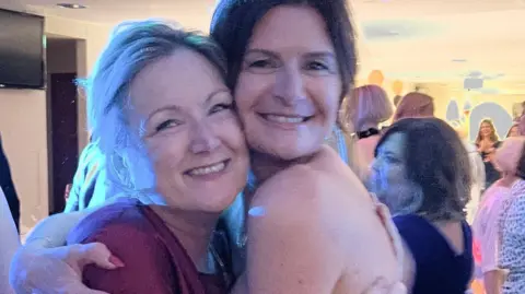 Jane Shorting and Leigh Lawson pictured at an event. They are cuddling each other and smiling at the camera. In the background there are lots of people in a dimly lit room.