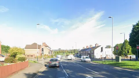 A Google streetview photo of a four-way junction. Three cars can be seen driving forward, there is a right turning lane as well as two lanes going in the opposite direction. There are houses ahead on both sides of the road.