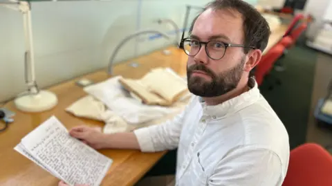 A bearded man with a white button down collar shirt at a desk holding a sheaf of paper.