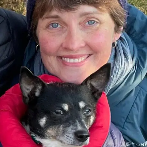 Paula Fraser A woman in a blue winter coat and woolly hat outdoors, holding a black and white dog in her arms.