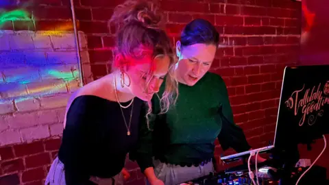 Supplied Two women look into a laptop, against a lit up brick wall. 