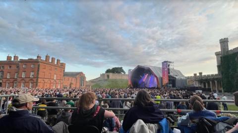 View from accessibility area at Kaiser Chiefs concert at Lincoln Castle with the backs of four people sat down looking across at the stage in the distance