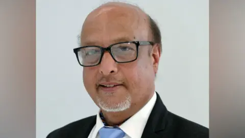 Slough Borough Council Head and shoulders official photo of Subash Mohindra wearing a blue tie and dark jacket. He has dark hair on the sides on his head and a grey goatee beard and wears dark framed glasses.