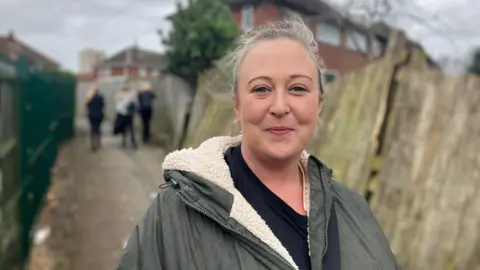 Elizabeth Baines/BBC A woman with blonde hair wears a green coat. It is shearling lined. Behind her, young people are blurred in a ginnel.