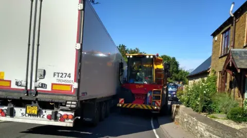 Farthinghoe Parish Council A white lorry struggles to get past a red lorry as it tries to drive around a corner in a village. 