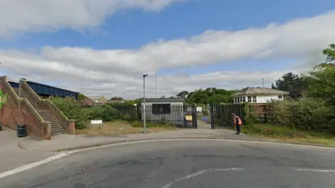 Google Brick steps up to footbridge over railway on left and man standing by black metal gates with buildings beyond.