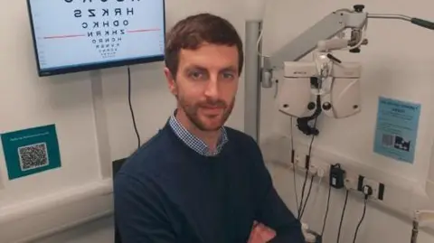 University of Bradford A man sits next to optician equipment and a screen with an eye test chart 