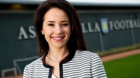 Birmingham 2022 A woman with dark hair and a black and white top is smiling at the camera. The Aston Villa club crest is blurred in the background.