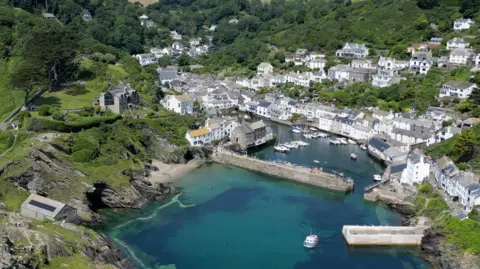 A shot of Polperro from the sky showing the entrance to the harbour and the two piers with lots of white cottages beyond and some properties on the hills around it