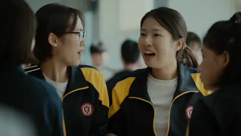 Four high school girls sit together in a cafeteria. They are wearing matching black-and-yellow athletic-school school jackets. Joo-in's friend, left, is turned slightly to the side wearing glasses, while Joo-in faces forward with her mouth open mid-expression 
