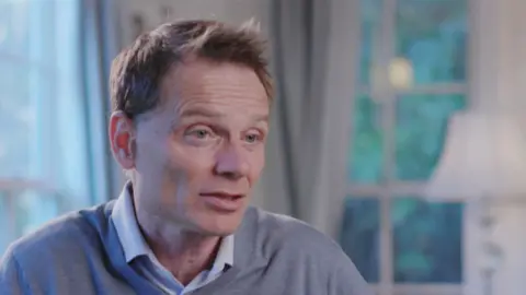 Professor Nicholas Bloom, a man with brown hair, wearing a V-neck jumper and collared shirt, is sitting in a room with a lamp and two corner windows, looking out at the trees. 