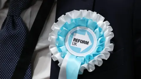 Close up of light blue Reform rosette on a man's suit. The man is wearing a white dotted dark blue tie. You cannot see the man's face. 