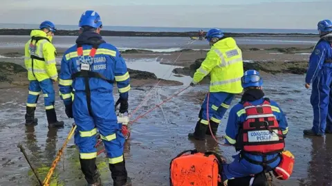 HM Coastguard Several workers in high-vis jackets on a muddy beach with winching materials helping rescue a walker