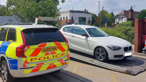 A police car parked next to a white car that is being towed away. There are buildings in the distance. It is a sunny day.
