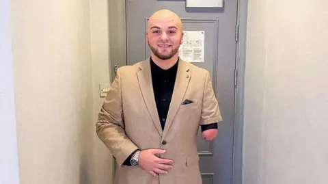 Ryan Rix Ryan Rix who is bald with a beard and one forearm, wearing a light brown suit, standing in front of a grey door