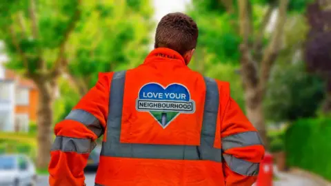 A man with his back to the camera is wearing a hi-vis orange jacket with a 'Love Your Neighbourhood' logo printed on the back.